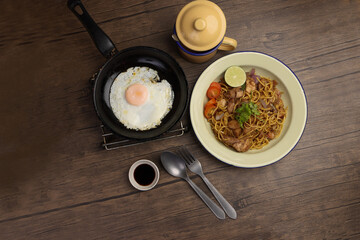 Stair fried yellow noodle traditional mee gorng tomato parsley lime on enamel metal plate cup sunny side up egg in frying pan fork spoon dark soya sauce