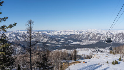 The funicular passes over a snow-covered valley. The cabs of the cable car are visible on the stretched ropes. A picturesque mountain range against a clear blue sky. Altai. Manzherok Ski Resort - obrazy, fototapety, plakaty