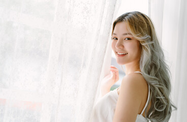 Obraz na płótnie Canvas Portrait of beautiful Asian girl hair coloring in blond standing near a window white curtains and turning to look and smile at camera. Alluring japanese woman posing near curtain in bedroom at home.