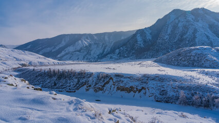 Fototapeta na wymiar A frozen river winds in a valley between steep banks. Fluffy hoarfrost on dry grass and on the ground. A picturesque mountain range against the blue sky. Altai