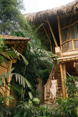 Beautiful slender young woman in a vintage dress against the backdrop of a bamboo house in a tropical garden. Eco house concept