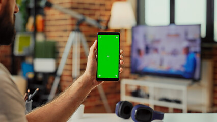 Young adult holding smartphone with green screen display in living room, using isolated template with chroma key and mockup. Analyzing blank copyspace background on mobile phone.