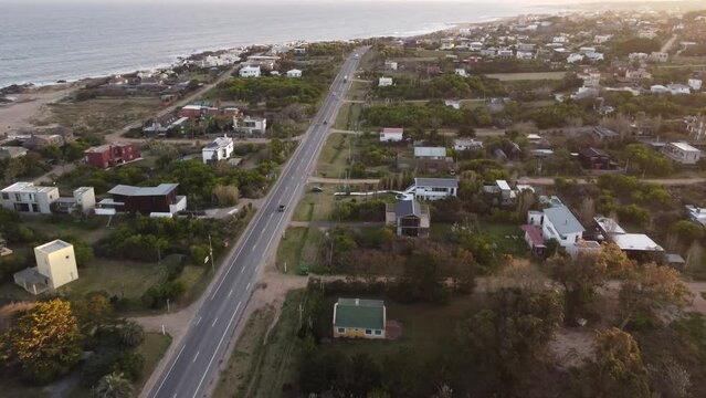 A dynamic aerial footage flying towards the sunset above the beautiful houses and traveling cars on the free road within the El Chorro area of Punta del Este Beach in Uruguay, South America. 