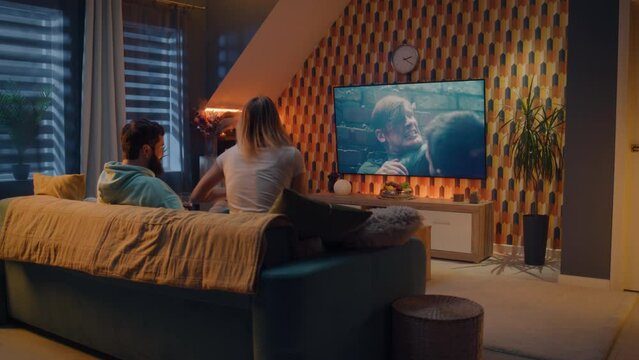 Woman jumping on couch. Couple on sofa in living room watching action movie on TV or criminal blockbuster on streaming service with snacks and chips, talking and discussing acting, resting at home.