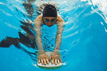From above of female swimmer in cap and glasses swimming with foam board in pool during training 