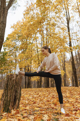 Full length of sporty woman in activewear
stretching legs near tree and doing exercises while leading heathy lifestyle and training in autumn forest  