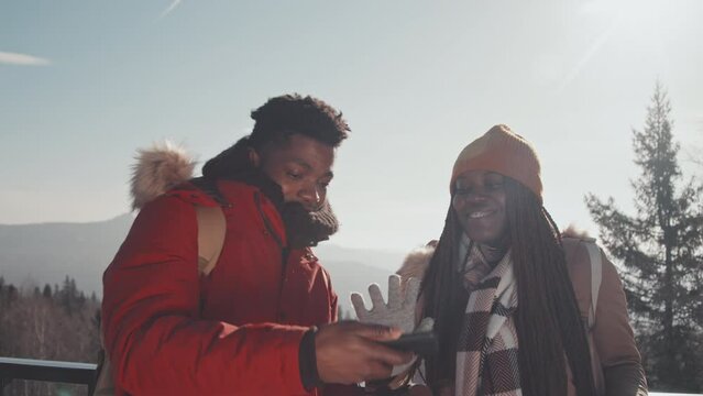 Modern young African American tourists standing at observation deck on mountain top on winter day taking selfie on smartphone camera