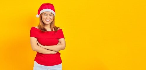 Portrait of smiling young Asian woman wearing christmas hat crossing hands on yellow background