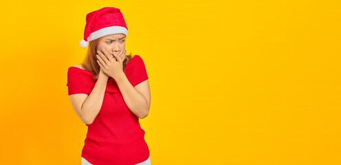 Surprised young Asian woman wearing Christmas hat covering mouth with hands on yellow background
