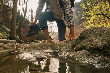 Male hiker crossing the stream barefooted walking by the creek with his shoes in hand during the...