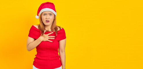 Fototapeta premium Shocked young Asian woman wearing Christmas hat and put hands chest on yellow background