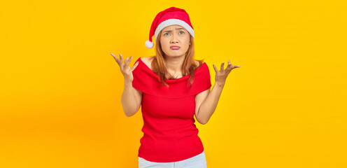 Fototapeta na wymiar Confused young woman wearing Christmas hat shrugging and hands on yellow background