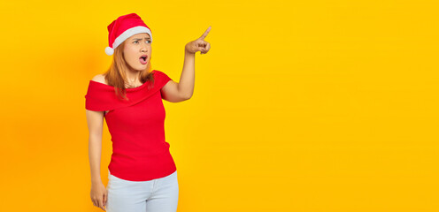 Surprised young Asian woman wearing Christmas hat and pointing sideways over yellow background