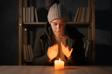 Freezing young woman in winter clothes warms her hands on lights with candles. suffers in no...