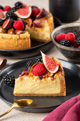 Vertical image of a piece of homemade baked cheesecake with fruits and berries: figs, strawberries and blackberries,  on a black plate.