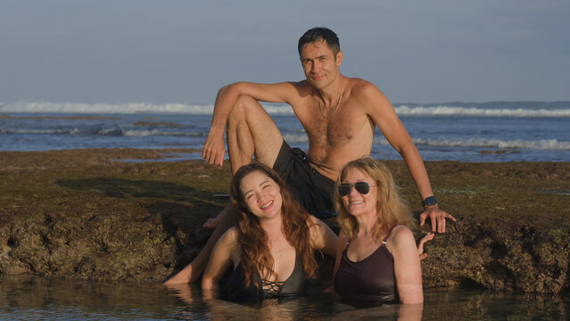 Handsome man sitting on a reef stone and two smiling women bathing in ocean water posing and smiling for the picture on beach at sunset. Happy time for a family spending time together. Soft focus.