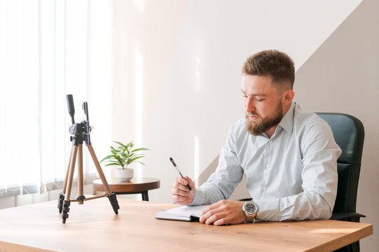 Cheerful bearded young man leading an online meeting using smartphone writing text information in notebook while sitting at table in an office in shirt. Positive guy talking on video link taking notes