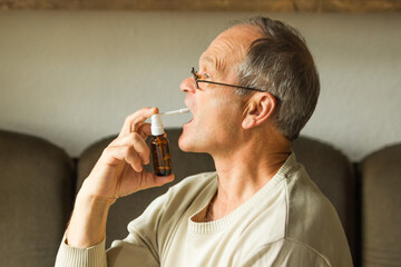 Sick caucasian senior man with open mouth using a throat spray against a dry cough, side view....