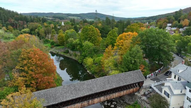 Rising aerial shot of covered bridge over river ascends to reveal mountainous pine forest with changing autumn foliage in the northeast. Quaint and scenic small town with main street and stream.
