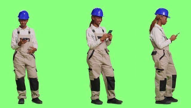 Female construction worker using smartphone app on full body green screen backdrop, surfing internet website online in studio. Contractor with overalls and hardhat using mobile phone.