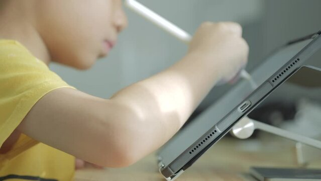 Asian boy writing a letter on a computer tablet with an electric pen in an online classroom due to Covid 19 pandemic. 5 years old kid studies at home.
