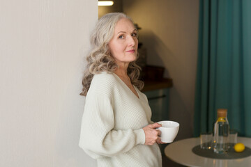 senior woman with gray hair  drinks tea  in the kitchen at home. concept without stress. mental health. Slow life. Enjoying the little things