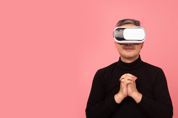 Portrait  of Asian man wearing a black turtleneck using virtual reality headset praying on pink background. Free from copy space. VR, future, gadgets, technology,  studying, video game concept