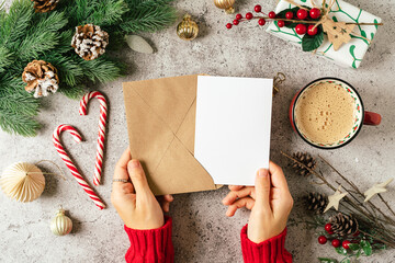 Desktop of woman hands in red sweater holding blank paper and envelope . Flat lay of gray background with cup of coffee and Christmas decoration. Top view mock up and copy space for text. - Powered by Adobe