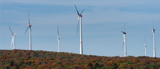 Colorful energy, wind turbines in a colorful autumn forest under a blue, slightly cloudy sky.