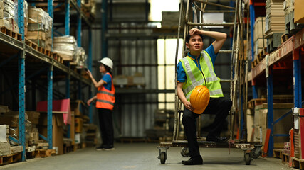 Tired male warehouse worker sitting and wipe the sweat away, taking break from hard work in large...