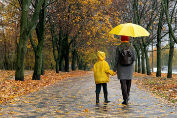 Woman with large yellow umbrella and child in yellow jacket are walking in the autumn park. Back view.