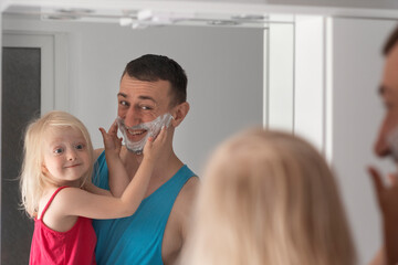Man with shaving foam on face holds little girl in arms. Dad and daughter have fun in bathroom by mirror