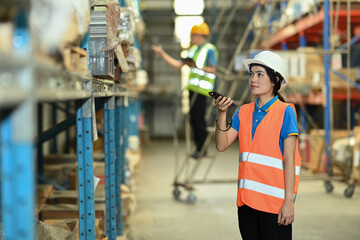 Fototapeta premium Female workers wearing hardhats and vests checking inventory boxes on shelf with barcode scanner
