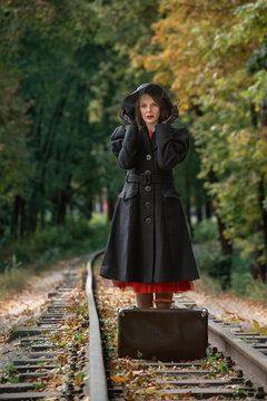 Middle-aged woman in retro black coat, broad-brim hat stands near vintage suitcase on railroad tracks. Vertical frame