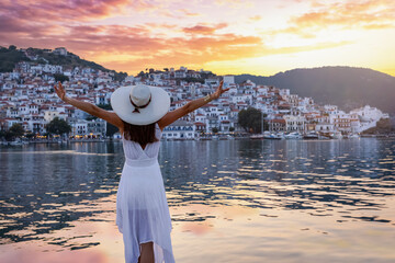 A happy tourist woman in a white dress looks at the idyllic town of Skopelos island, Spordes,...