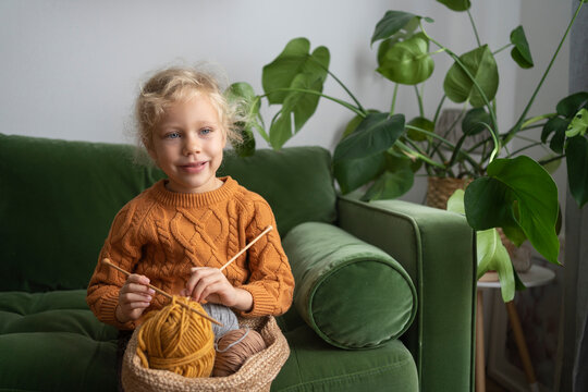 Girl holding basket with balls of wool and knitting needles on sofa at home