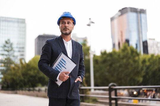 Thoughtful engineer wearing hardhat standing at footpath