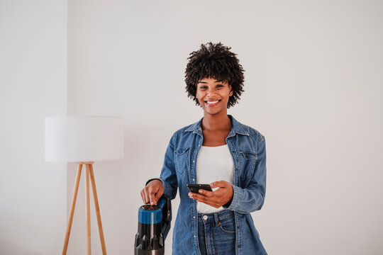 Happy woman standing with vacuum cleaner and smart phone in front of wall at home