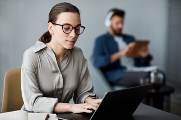 Young serious brunette woman in smart casualwear and eyeglasses sitting in front of laptop and typing while carrying out assignment