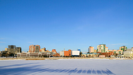 Fototapeta premium Panorama of a frozen river and buildings illuminated by the sun on a winter day against a blue sky