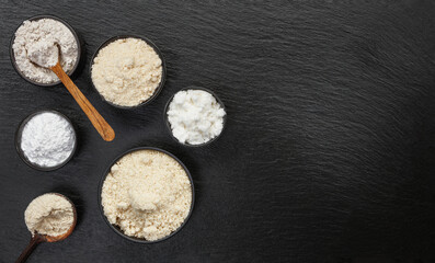 Bowls and wooden spoons with different types of flour on a black slate background, top view. Space...