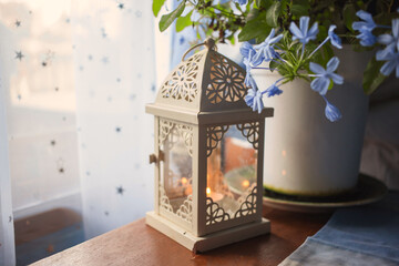 Ornate vintage style lantern with burning candle inside standing near blooming blue flower pot on...