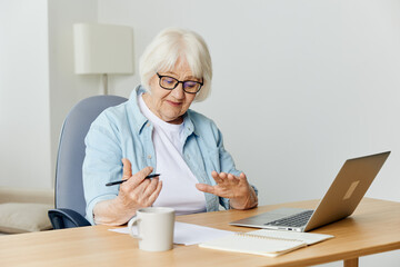 a pleasant, stylish elderly woman is sitting at home in a bright interior and working on a laptop makes notes on a piece of paper. The concept of working from home