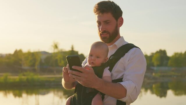 Close up young handsome father with a little newborn baby in a slings carrier. Use phone typing smiling on street. Ergo backpack. Parents. Gadget. Smartphone. Cell. Slow motion