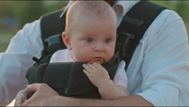 Beautiful portrait of a newborn baby looking around in a slings carrier. Young father holding a child on street. Ergo backpack. Parenthood. Dad. City.Slow motion