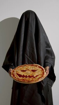Vertical studio shot of unrecognizable young woman in black ghost costume holding pizza with jack o lantern face for Halloween partyVertical studio shot of unrecognizable young woman in black ghost co