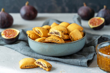 Biscuits filled with fig jam on grey bowl, fresh figs, jam in jar, napkin on grey concrete background. Close up. 