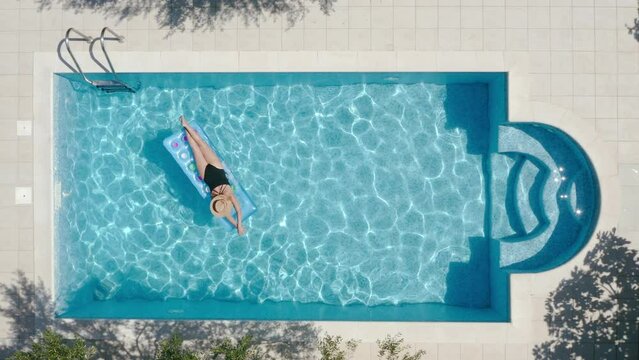 Top view swimming pool with relaxing woman in black swimsuit, straw hat drifting on inflatable mattress on the blue ripples water surface under olive trees. Holidays, recreation 4K video concept.
