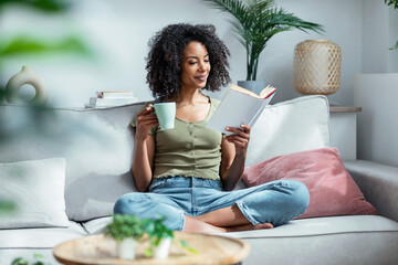 Pretty woman reading a book while drinking a cup of coffee sitting on sofa at home. © nenetus