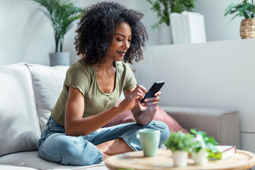 Shot of confident woman using her mobile phone while drinking coffee sitting on sofa at home.